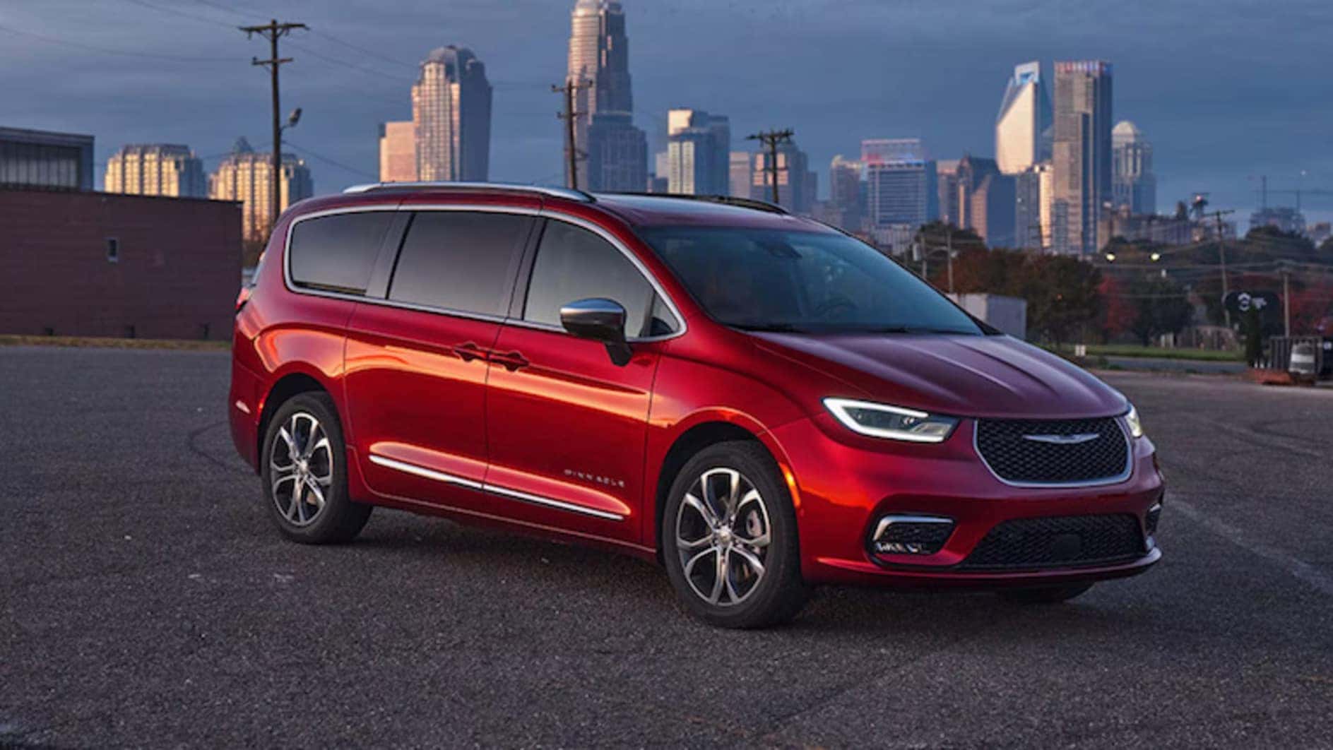 A red 2026 Chrysler Pacifica minivan parked in a parking lot, with a city skyline visible in the distance