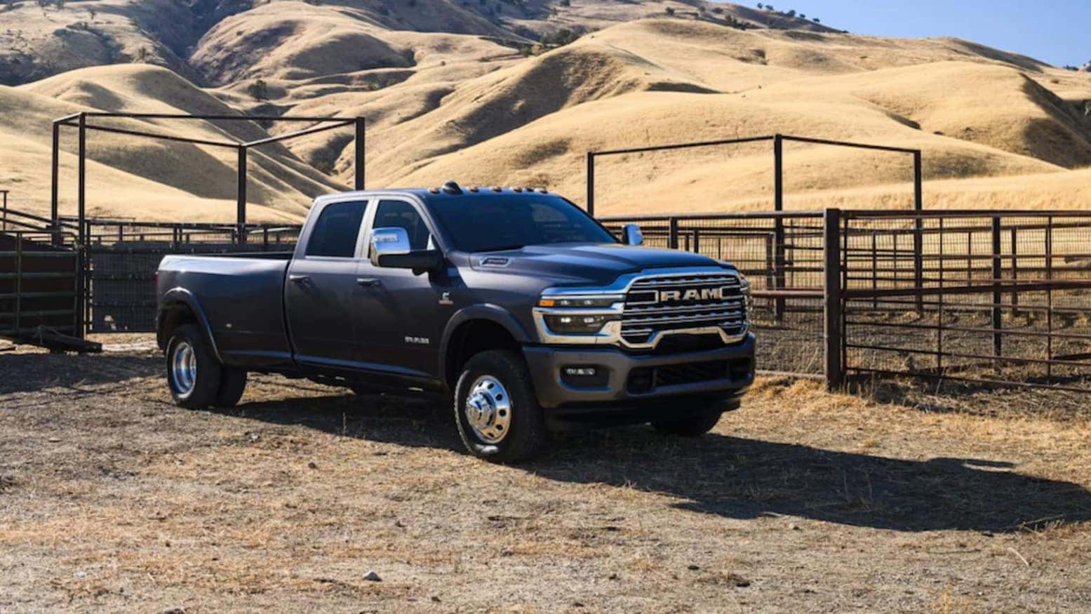 A dark-gray 2026 Ram 3500 truck parked in front a fenced-in pasture with hills in the background