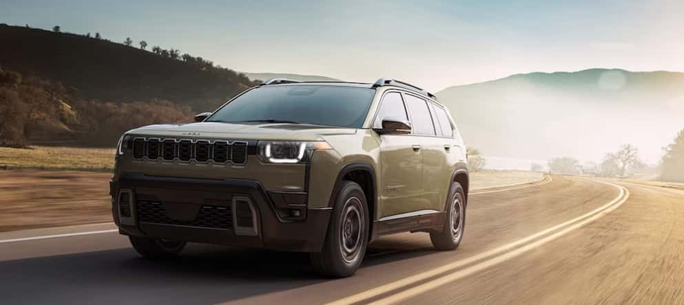 An olive green 2026 Jeep Cherokee SUV driving down a misty road with mountains in the background