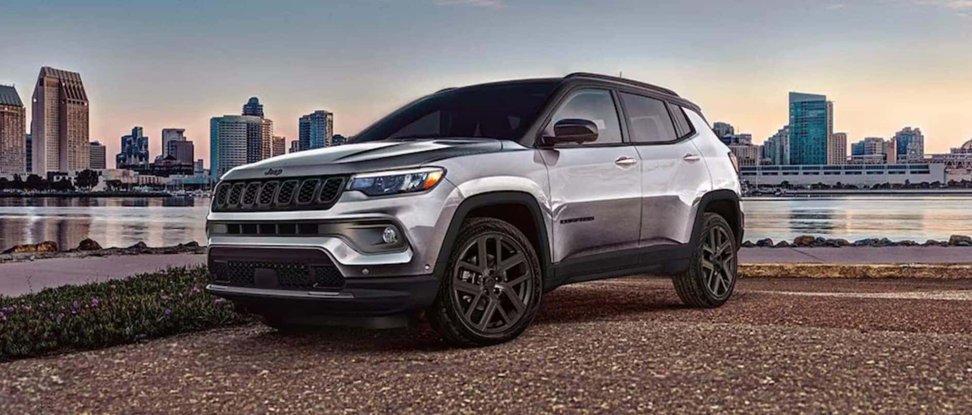 A silver 2026 Jeep Compass parked in front of a body of water, with a city skyline in the distance