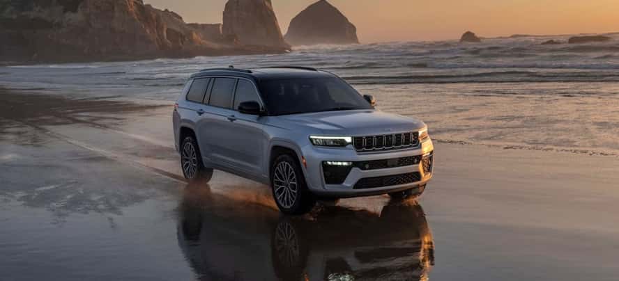 A gray 2026 Jeep Grand Cherokee SUV driving down a beach at sunset