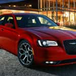 A red Chrysler 300 sedan parked at night, with modern architecture and lighting in the background, highlighting the vehicle's sleek design and distinctive grille.