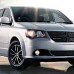 Silver Dodge Grand Caravan parked on a wet pavement with sunlight reflecting off its hood, showcasing its front grille and headlights.