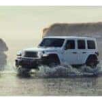 sideview of white jeep wrangler driving on beach in shallow water