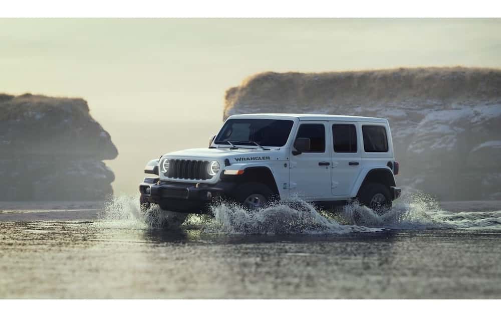 sideview of white jeep wrangler driving on beach in shallow water