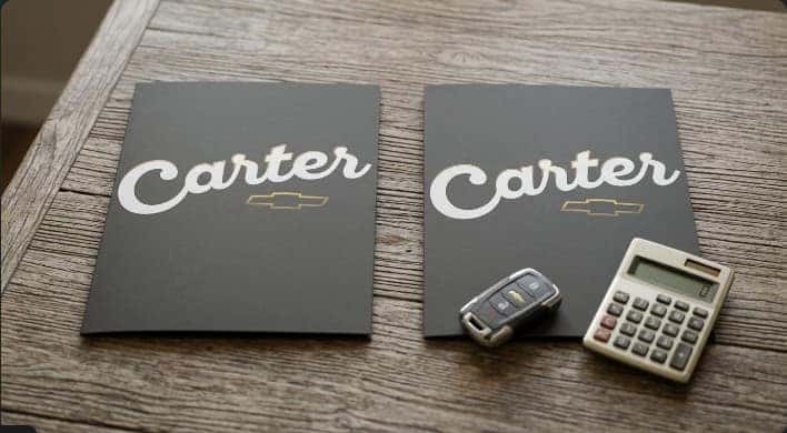 Flat lay of a car key fob, calculator, and two Carter Chevrolet finance folders on a wood table, representing comparing auto loan offers