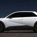 Side view of a white Hyundai IONIQ 5 XRT parked under a starry night sky, showcasing its angular futuristic design, black wheels, and rugged lower body cladding against a desert backdrop.