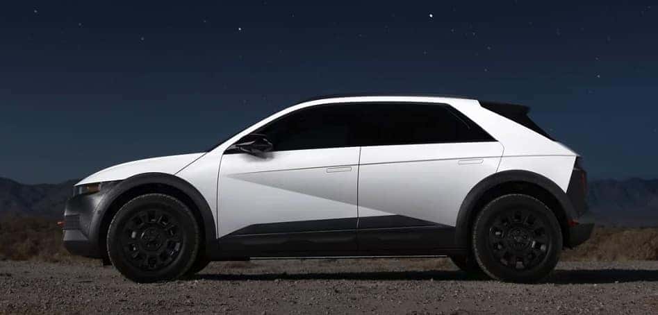Side view of a white Hyundai IONIQ 5 XRT parked under a starry night sky, showcasing its angular futuristic design, black wheels, and rugged lower body cladding against a desert backdrop.