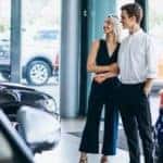 A couple is standing in front of a large window looking at cars.