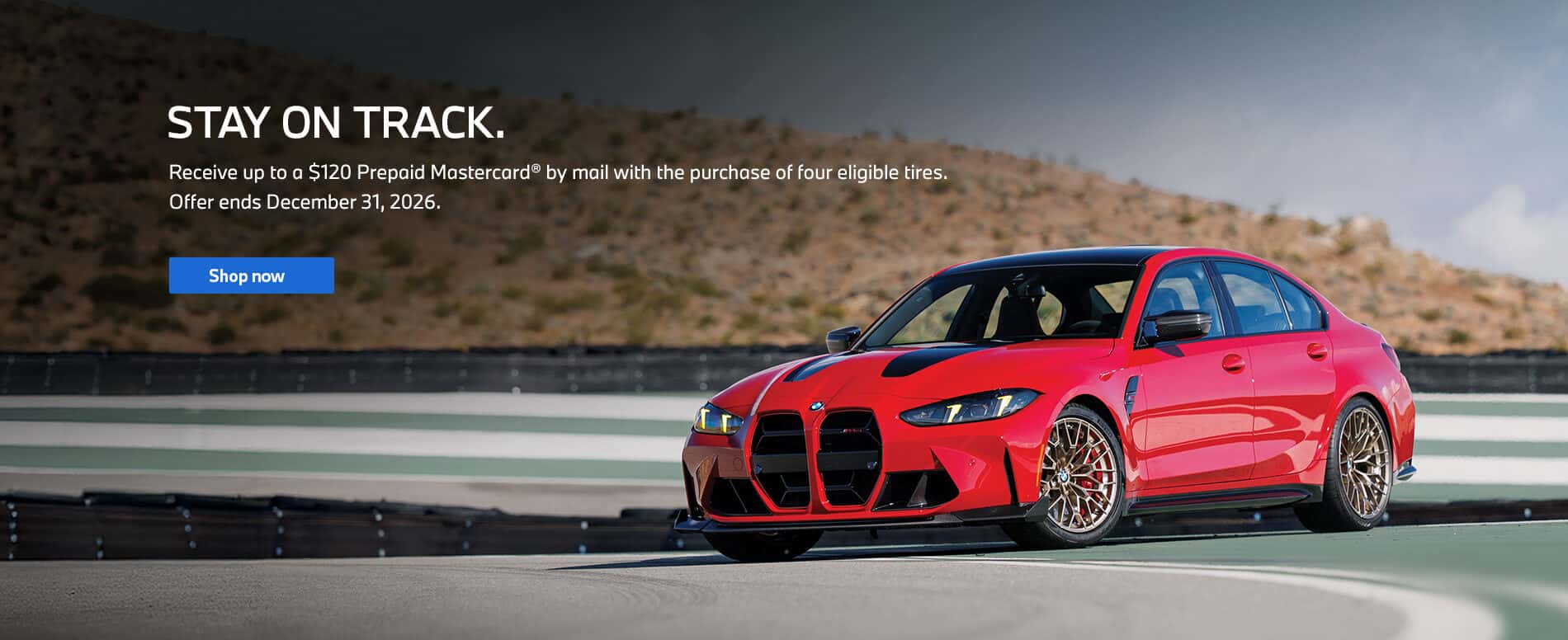Red BMW driving around a racetrack curve, painted with green and white stripes. A desert hill and a cloudy skyline the background.
