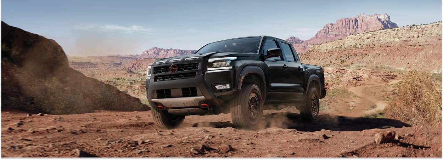 Black 2026 Nissan Frontier pickup truck driving on a dirt trail in front three-quarter view.