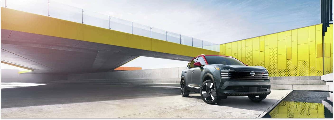 Two-tone gray and red 2026 Nissan Kicks parked in front three-quarter view near a modern yellow structure.