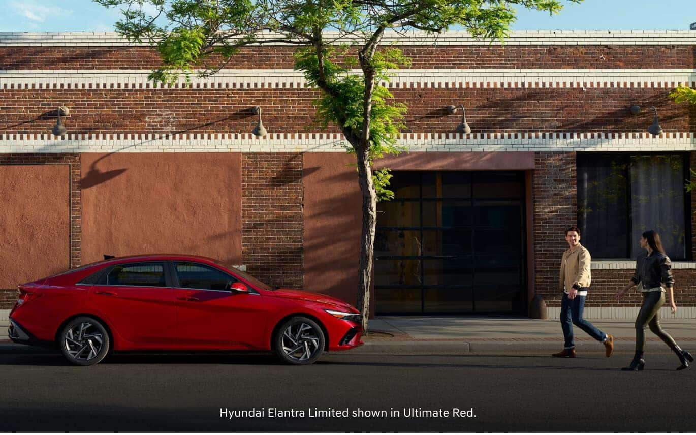 Side profile view of a man and a woman walking past a 2026 Hyundai Elantra Limited in Ultimate Red parked on a city street.