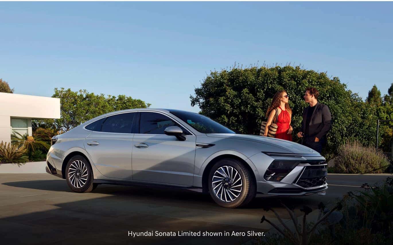 Side profile view of a woman in a red dress and a man in a black jacket standing near a 2026 Hyundai Sonata Limited in Aero Silver.