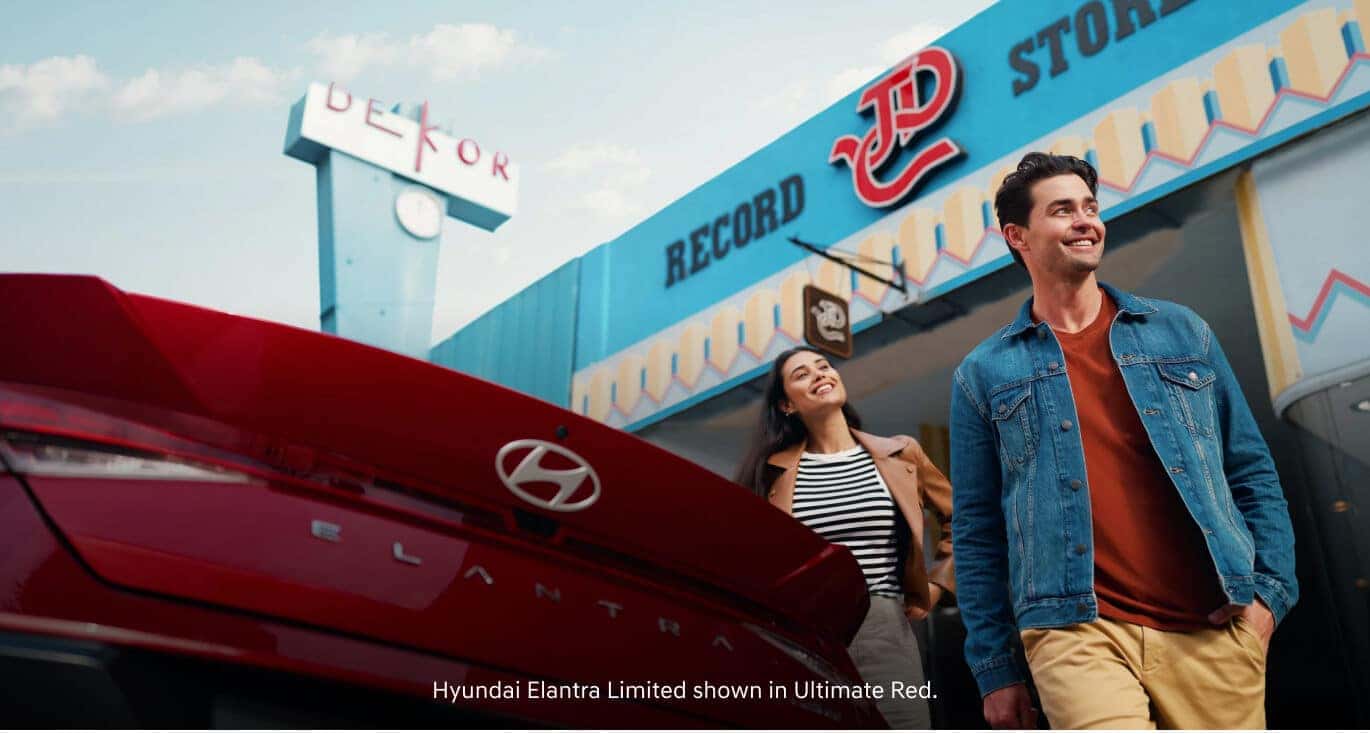 Rear view of a smiling man and woman walking past a 2026 Hyundai Elantra Limited in Ultimate Red outside a record store.