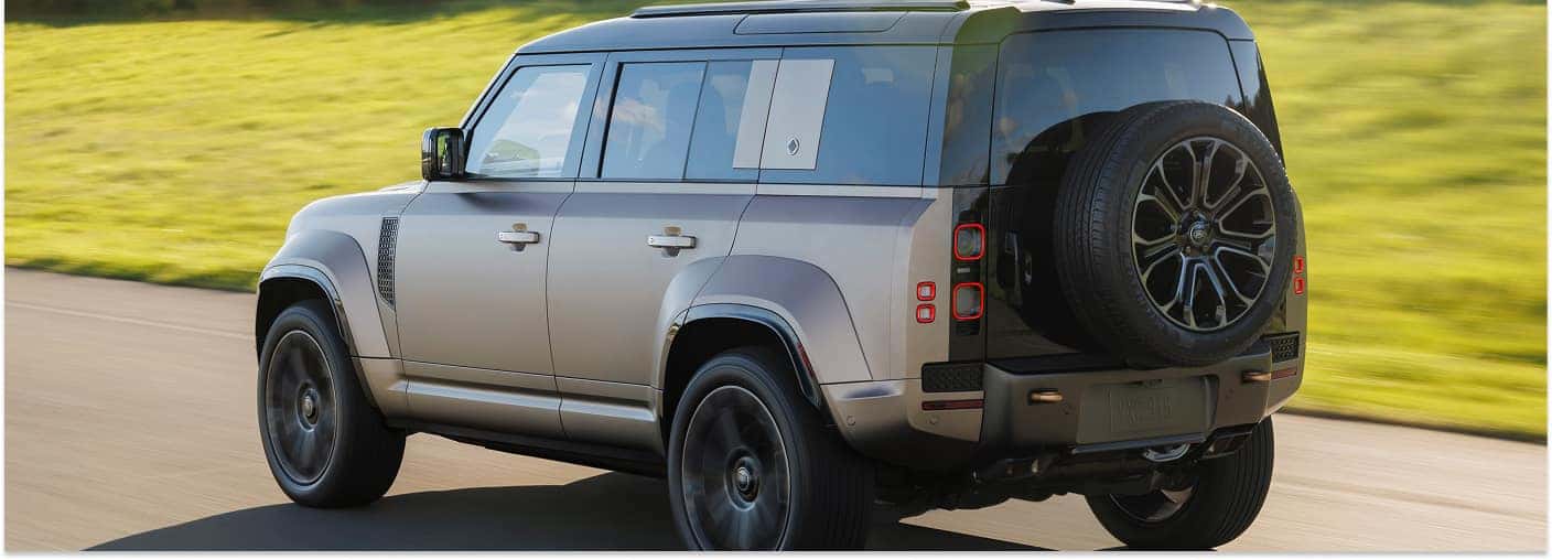 A gray 2025 Defender SUV drives along an asphalt road, showcasing its rear profile with a two-tone black roof and a rear-mounted spare tire against a blurred green background.