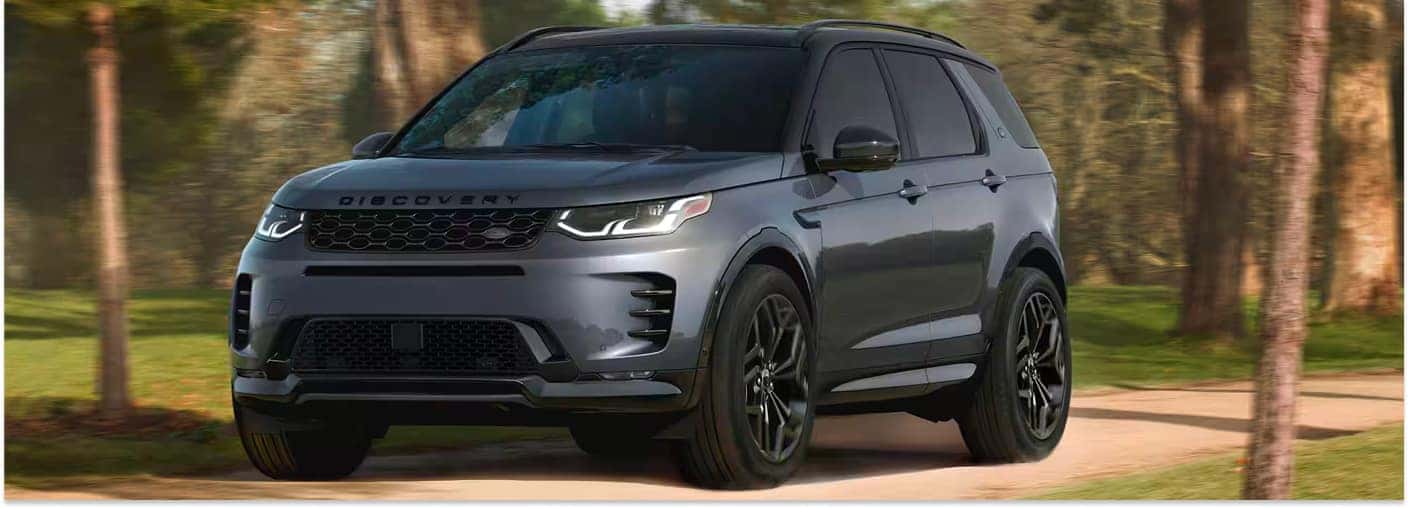 A front three-quarter view of a dark gray 2026 Discovery Sport driving on a path through a sunlit, wooded area.