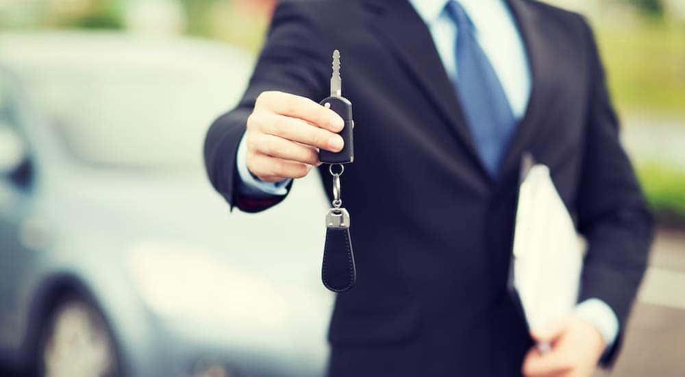 A salesman in a suit is holding out a car key and holding paperwork.