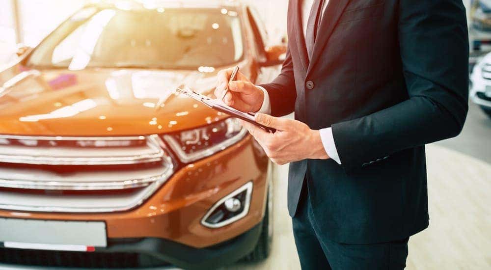 A car salesman is standing in front of an orange car inside a dealership showroom and holding a clipboard.