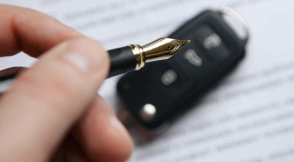 A closeup shows a hand holding a pen over a car key and paperwork,