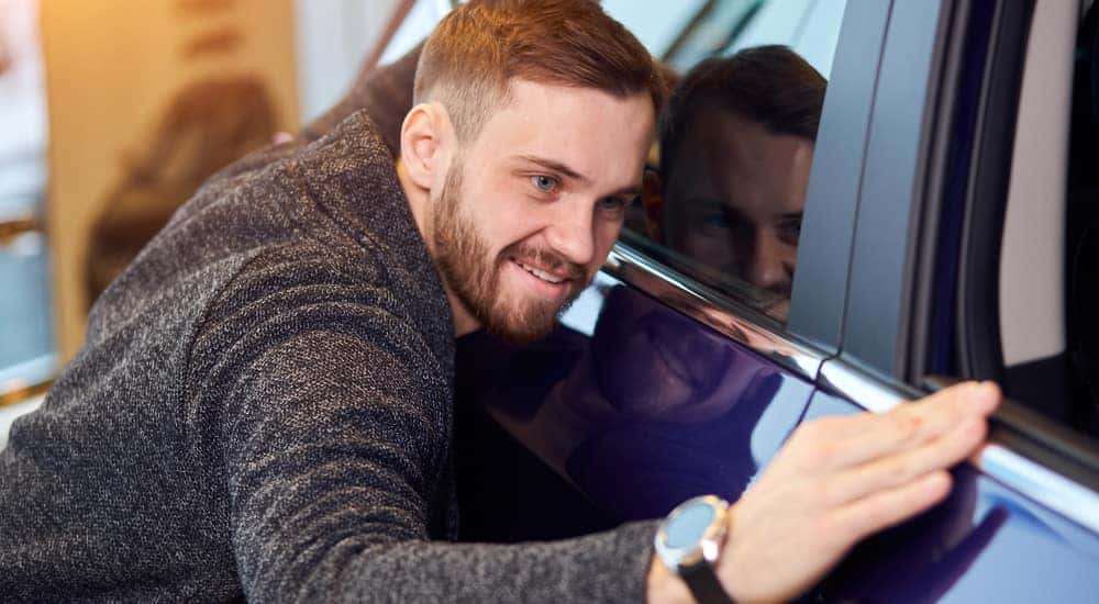 Man in a grey sweater, smiling with his arms around a blue car