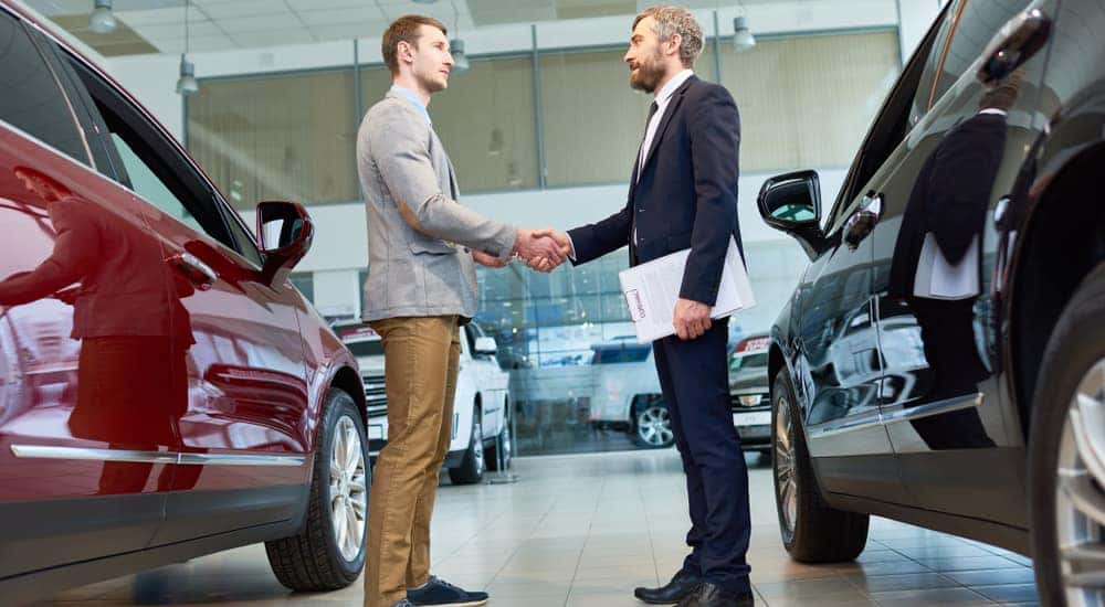 Man in a grey blazer and brown pants shaking hands with a man in a black suit in between a red and a black vehicle