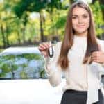Woman in a white blouse and black skirt giving a thumbs up and holding up a car key in front of a silver sedan