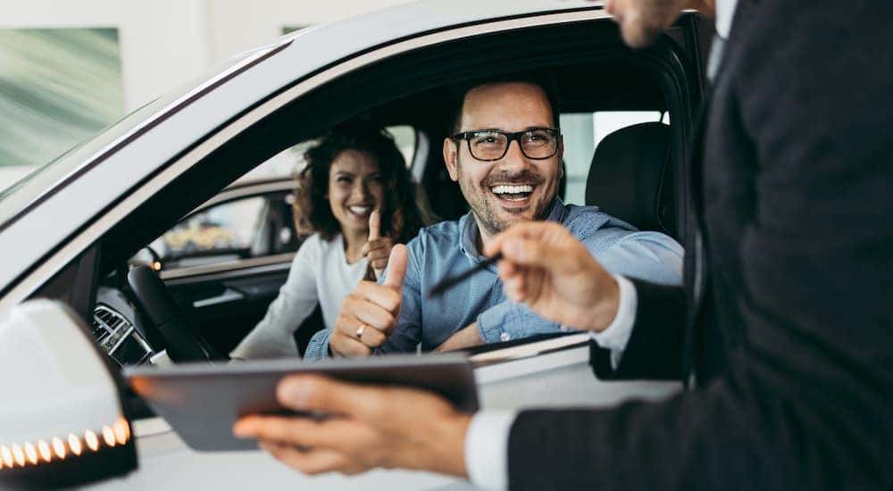 A couple is shown smiling from the front seat of a car while talking to a salesperson.