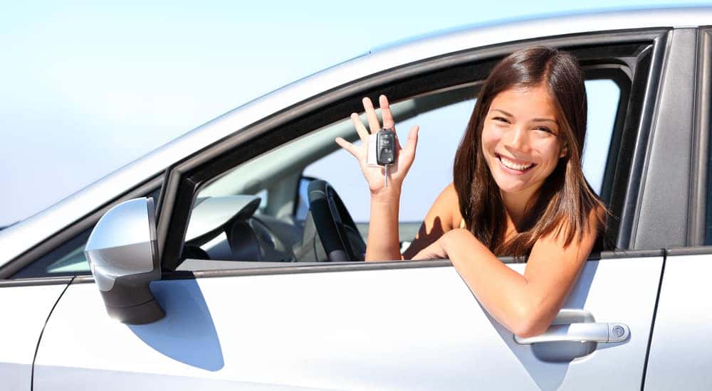 Girl with brown hair sitting in the driver's seat of a silver car and holding up a car key outside the window