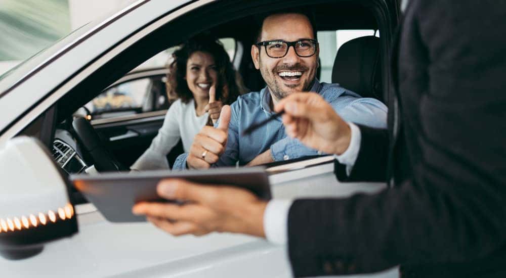 Man in a blue shirt and glasses sitting in driver's side of vehicle next to woman, giving thumbs up to a man with a tablet outside the car.