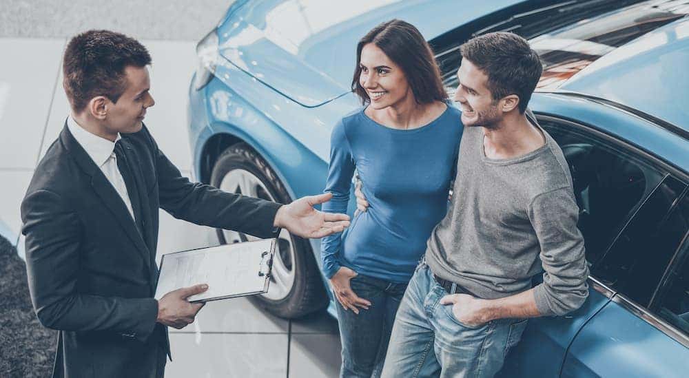 Two people are shown talking to a salesperson in front of a blue car.