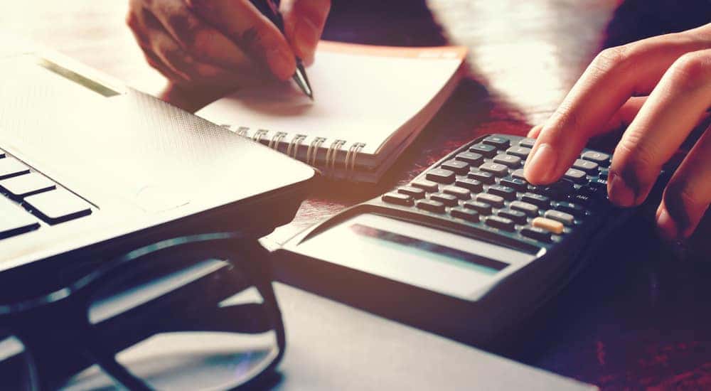 Hands using a calculator on a desk next to a pad of paper, a laptop, and eyeglasses