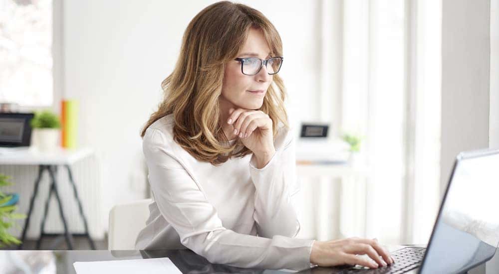 Woman in a white shirt sitting at a black desk with a laptop, smartphone, paper, and pen