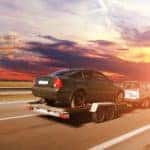 A small silver SUV is towing a used car on a trailer towards a sun set near Cincinnati, OH.