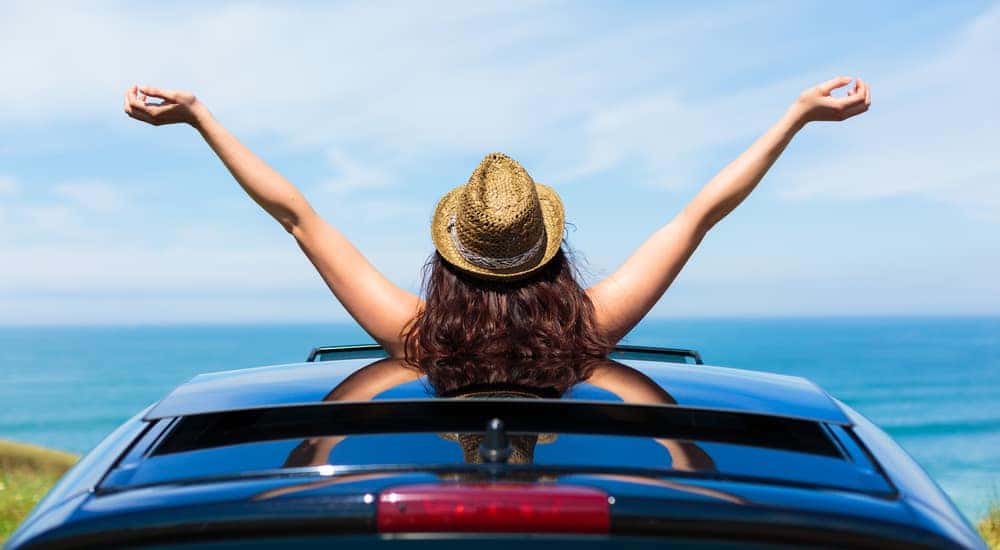 Woman with brown hair and a light brown hat out of the sunroof of a black sedan with her arms up
