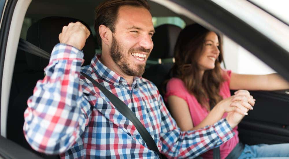 Woman in a pink shirt sitting in the driver's seat of a vehicle, holding hands with a man in a plaid shirt with his arm up and smiling