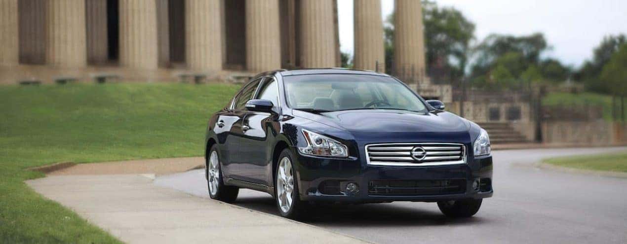 A blue 2012 used Nissan Maxima is shown in front of a public building with columns.