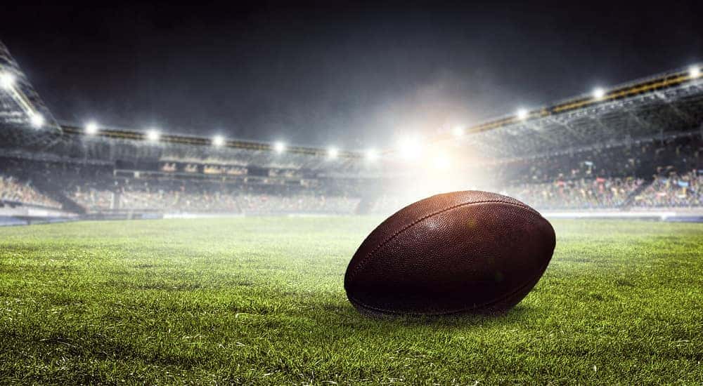 Brown football sitting on a green, grassy football field surrounded by fan seating and lights