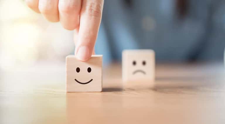 One wooden block with a black smiley face on it being pushed forward on a table from next to a cube block with a frowning face on it by one single finger
