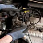 Man's hands in blue gloves holding a thick black belt above the open hood of a vehicle