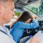 Man writing on a driver's test evaluation sheet while a girl sits in the front seat of a vehicle, upset with her head on the steering wheel
