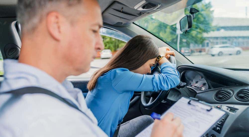 Man writing on a driver's test evaluation sheet while a girl sits in the front seat of a vehicle, upset with her head on the steering wheel