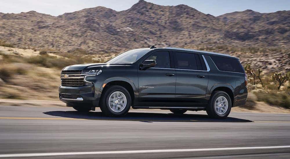 A grey 2021 Chevy Suburban is shown from the side driving on an open road after leaving a used car dealership/