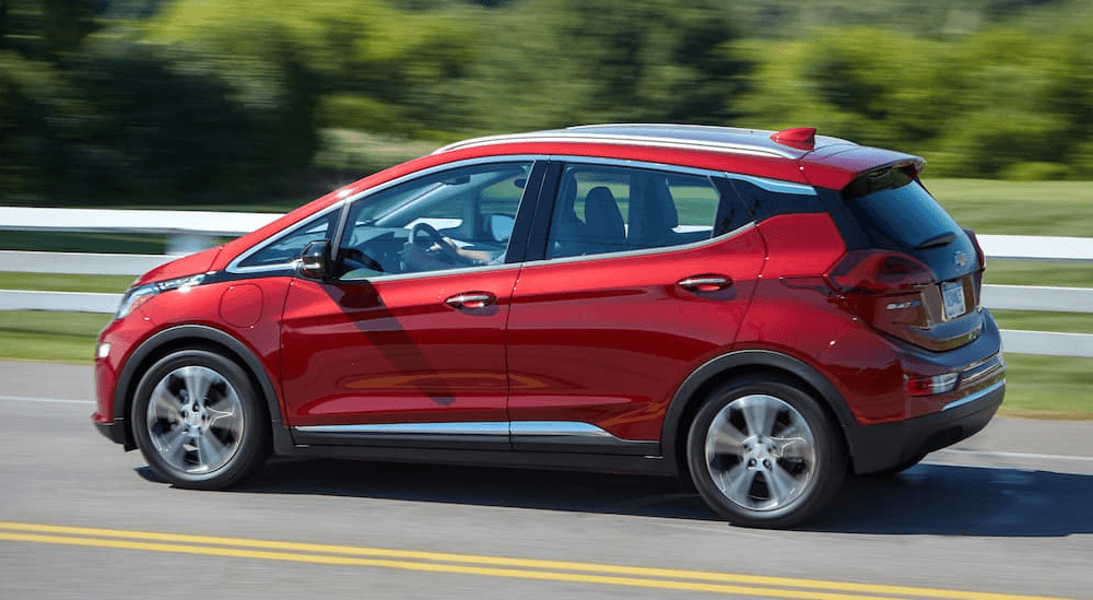 A red 2020 Chevy Bolt EV is shown from the side driving next to a white fence and field.