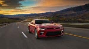 A red 2017 Chevy Camaro SS is driving on a highway in front of a vibrant orange and teal sunset.