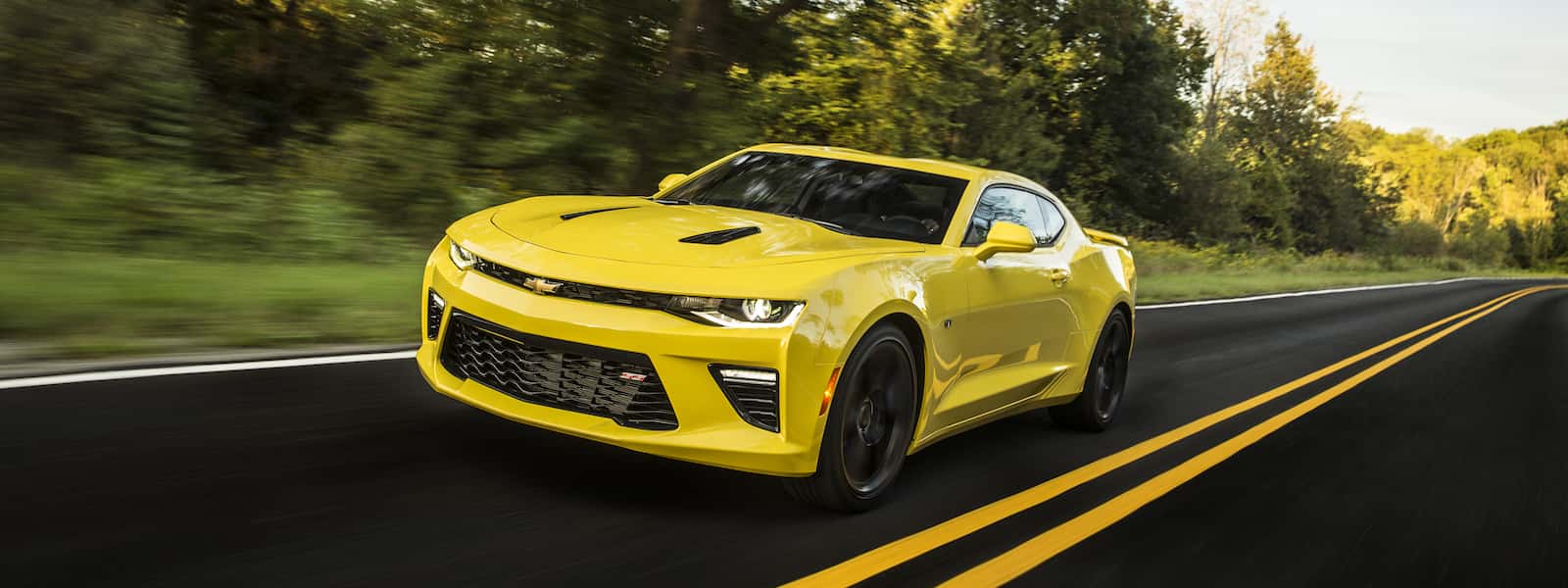 A yellow 2016 Chevy Camaro driving down a scenic road with trees in the background.