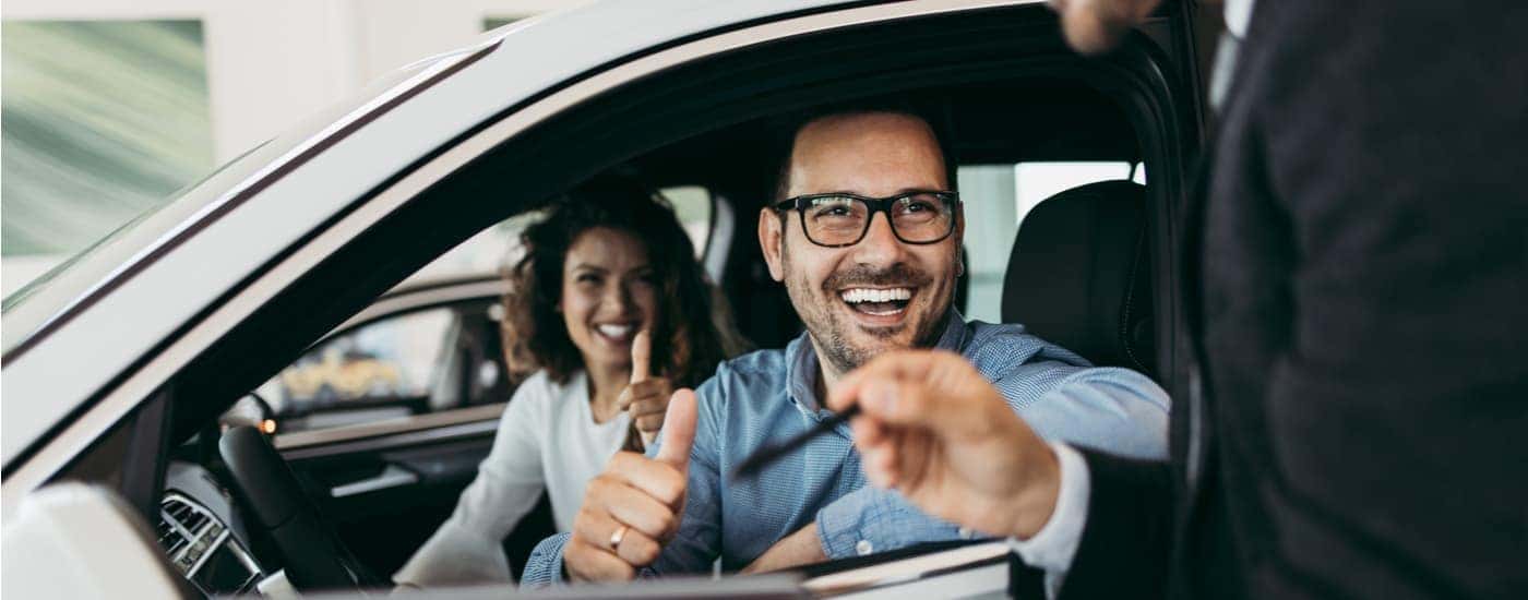 A salesman is shown speaking to customers sitting inside their new vehicle at a car dealership.