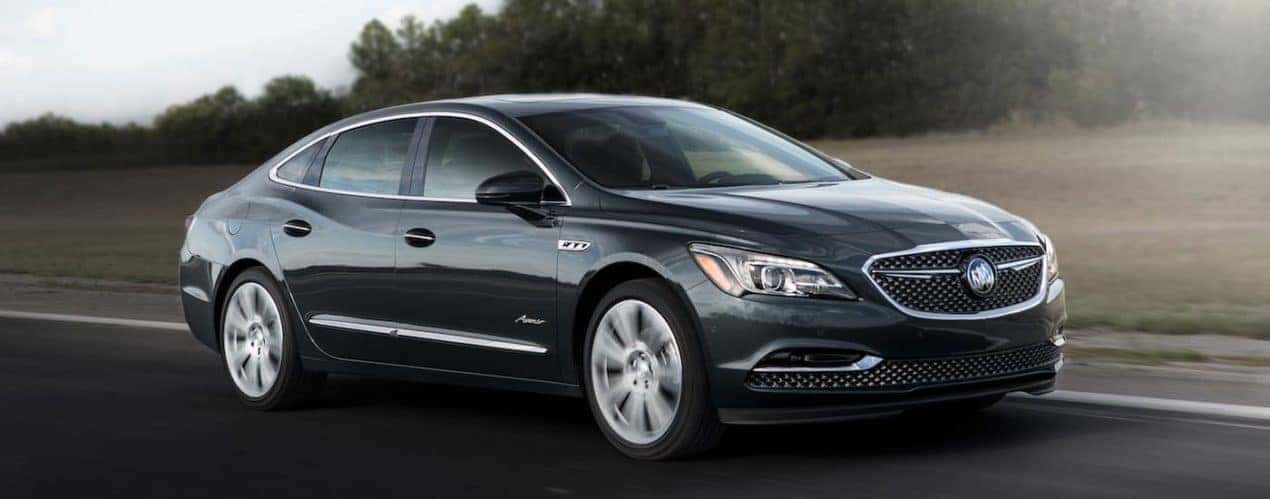 A black 2018 used Buick LaCrosse Avenir is shown driving by grassy field and trees.
