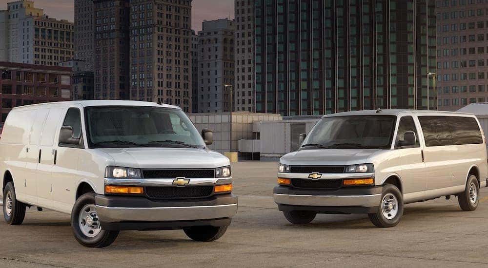 A white and a silver Chevy van are parked in an empty city lot.