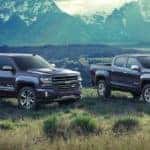 A dark grey 2018 Chevy Silverado and Colorado are parked on grass in front of distant mountains.